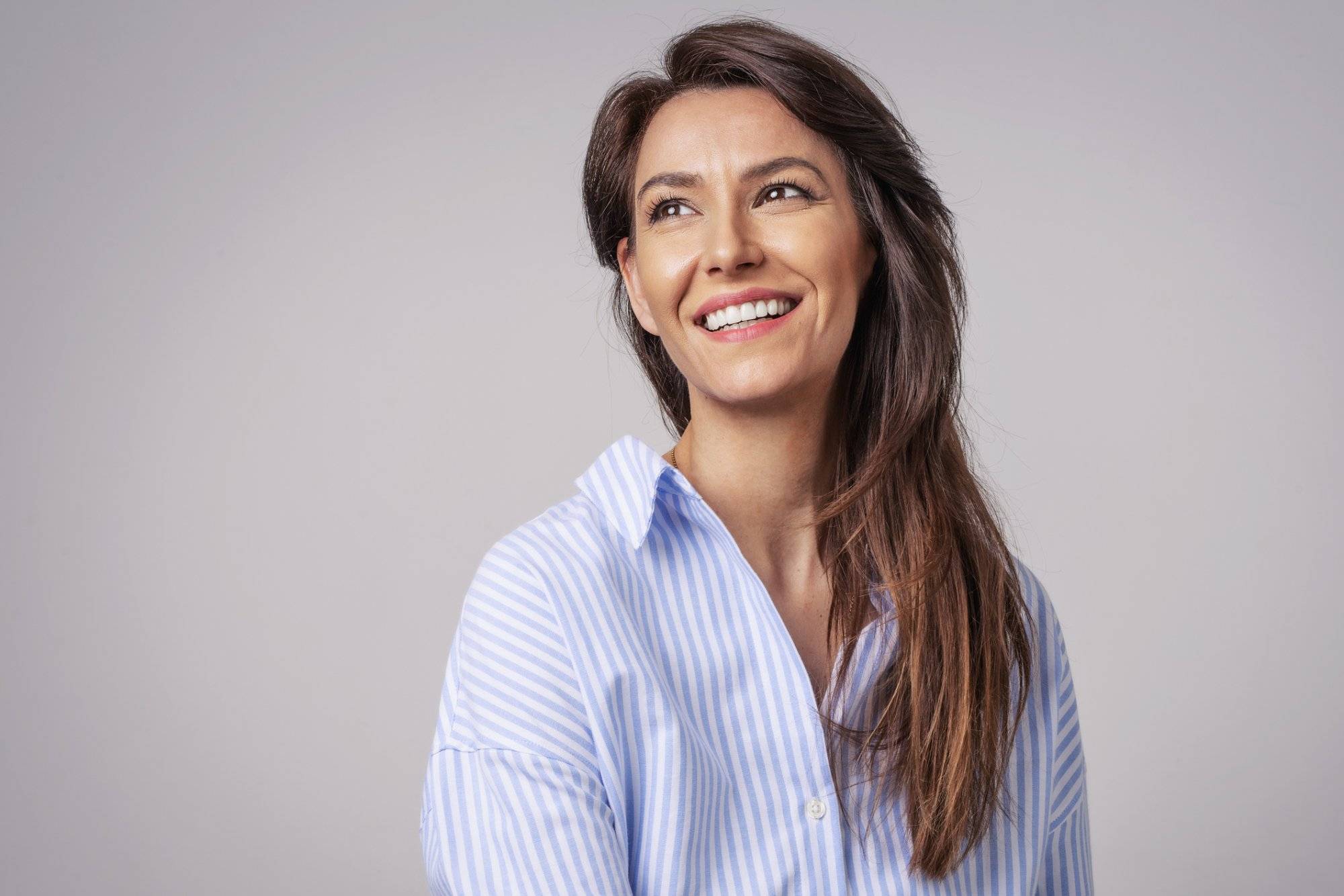 Smiling brunette businesswoman sitting against gray background. Confident female professional is wearing blue shirt. She is having brown hair. Copy space.